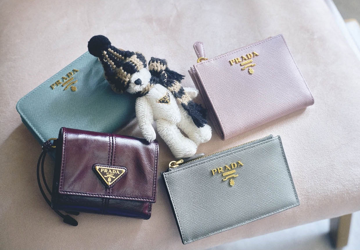 上段右から、「サフィアーノ」レザーはキズや汚れがつきにくく、長くきれいに使えるのが嬉しい。レタリングロゴとスモーキーなピンクが大人っぽくて上品。薄くてコンパクトな二つ折り。ウォレット［H8.5×W 11.5×D2、カード入れ8］￥103,400、ポンポン付きのニットキャップとマフラーを巻いたほっこりスタイルが可愛い。チャーム￥116,600、レトロシックなグリーン。使い勝手のいい二つ折り。ウォレット［H8.5×W12×D3.5、カード入れ6］￥138,600、下段右から、クールなブルーがスタイリッシュ。フラグメントケース［H8×W13、カード入れ5］￥88,000、柔らかくてなじんだレザーの質感とトライアングルロゴがかっこいい。トレンド感のあるボルドー。ウォレット［H8.5×W10×D2、カード入れ4］￥108,900（すべてプラダ／プラダ クライアントサービス）※すべて予定価格